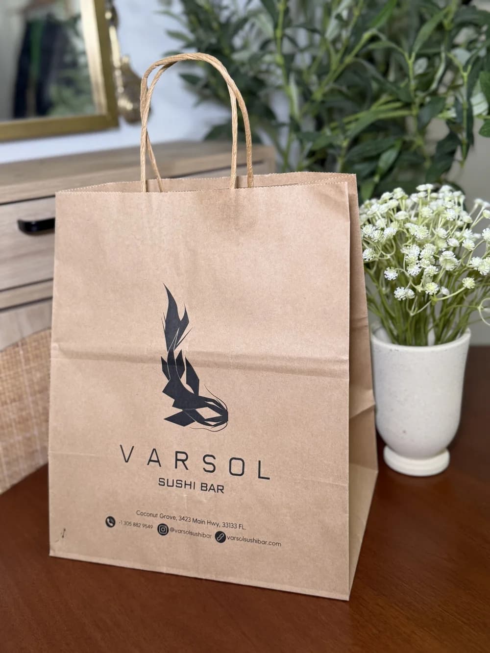 A kraft paper takeout bag with twisted handles printed with the Varsol Sushi Bar logo, photographed on a wooden table beside a vase of white flowers.