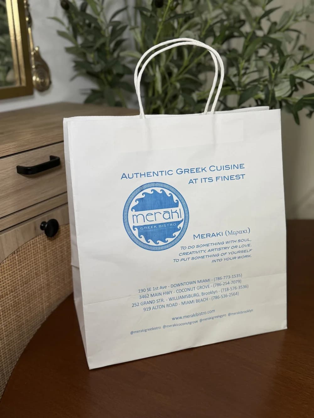 A white paper takeout bag with twisted handles, printed in blue with the Meraki Greek Bistro logo and location details, photographed on a wooden table with greenery behind.