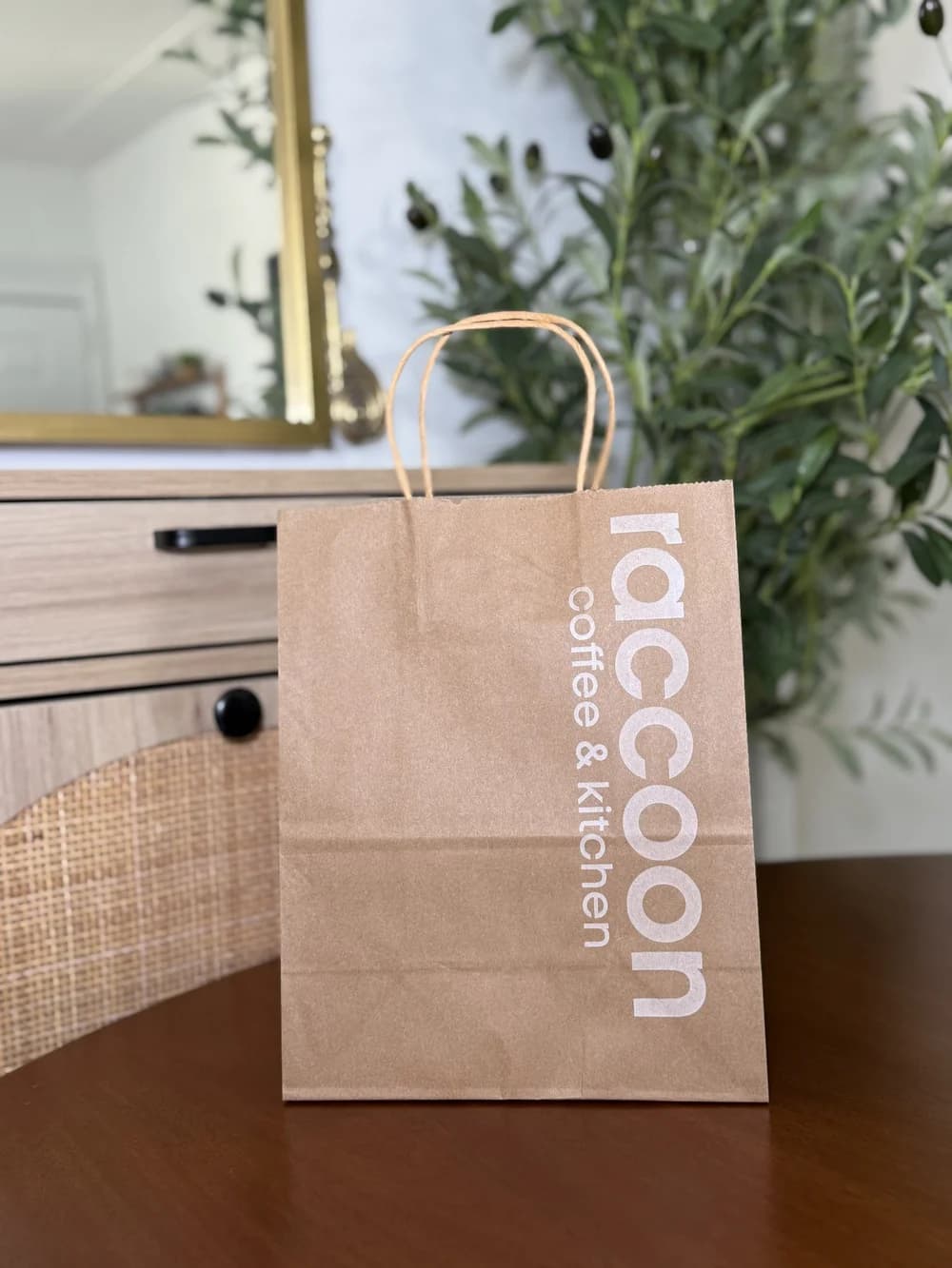 A kraft paper takeout bag with twisted handles printed with a vertical white "raccoon coffee & kitchen" logo, staged on a wooden table with greenery and a mirror behind.