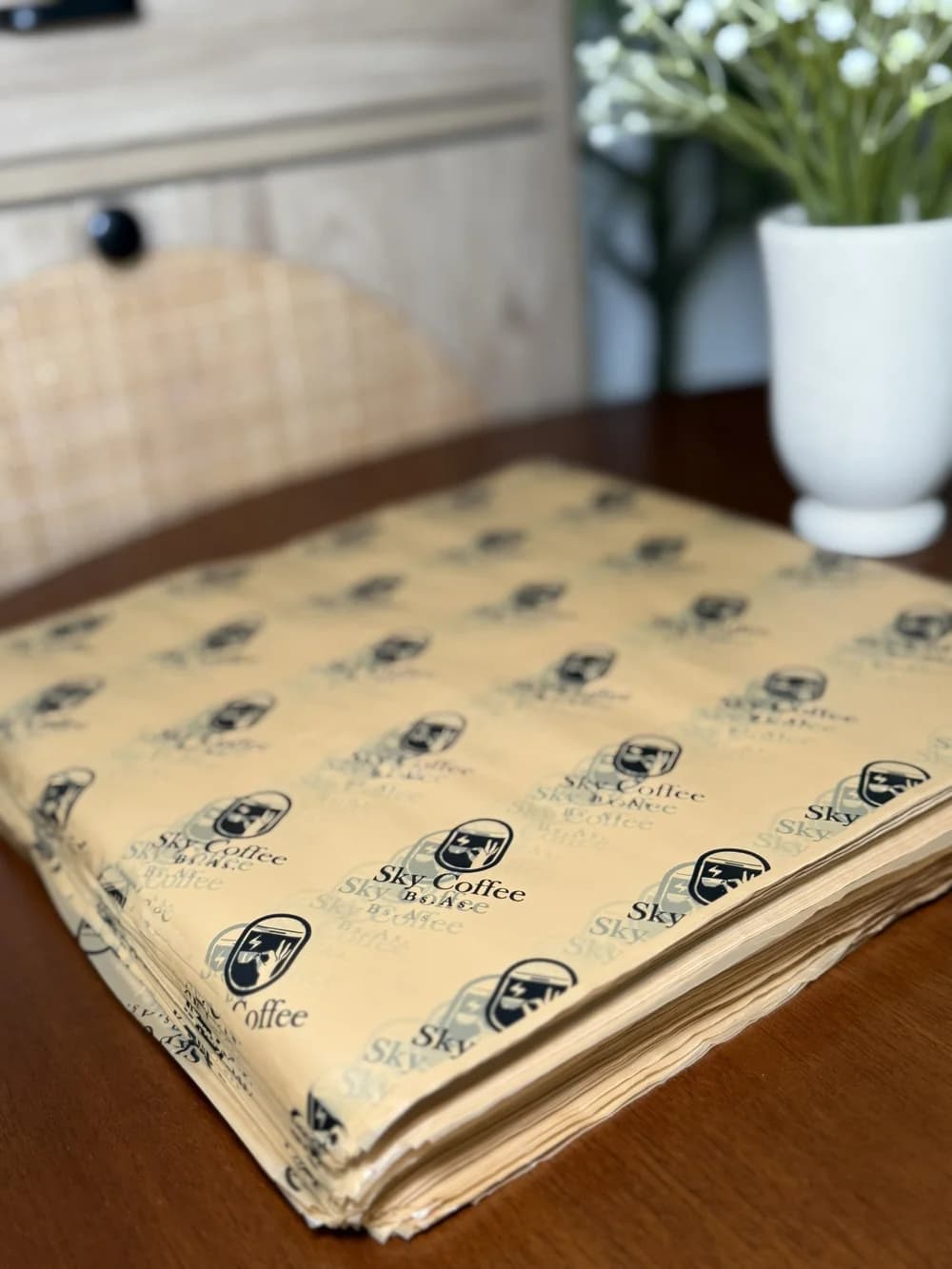 A stack of custom-printed food-grade tissue wrapping paper sheets with a repeating "Sky Coffee" logo pattern, photographed on a wooden table beside a vase of flowers.