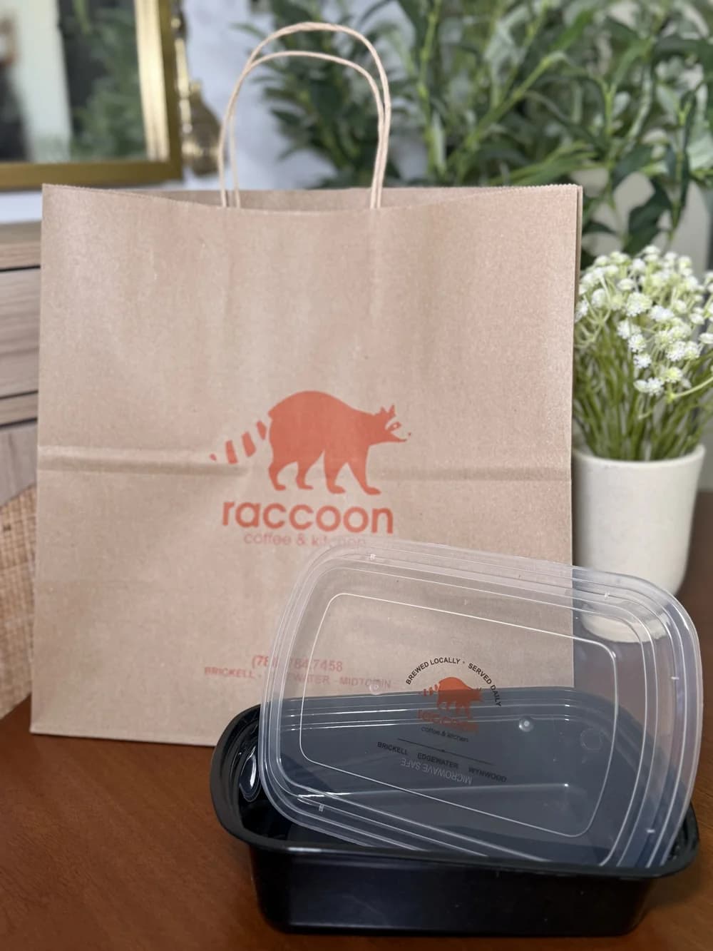 A kraft paper takeout bag with twisted handles printed with the Raccoon Coffee & Kitchen logo, photographed on a wood table beside a matching black microwave-safe food container with printed clear lid.